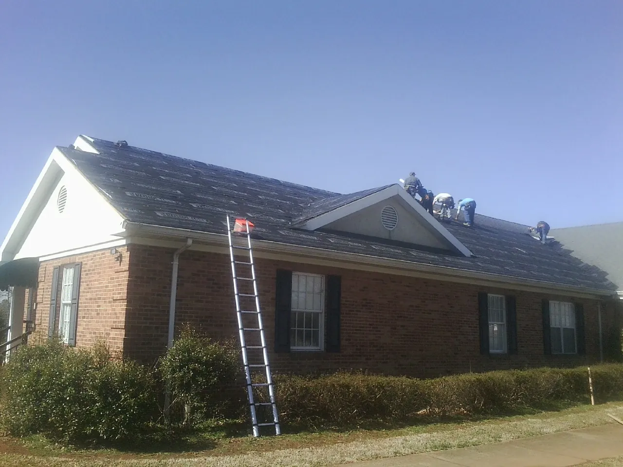 Crew tearing shingles off a brick ranch roof with a ladder set — Emergency Roof Repair work by our crew in Sale City