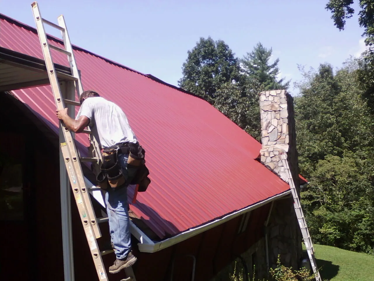 Roofer on a ladder beside a red metal roof and stone chimney — Soffit & Fascia Repair by Roofing Brothers in Sale City